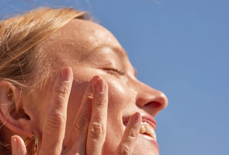 A person is applying sunscreen to their face while smiling under a clear blue sky. The sunscreen tube in their hand is labeled "kinfield.
