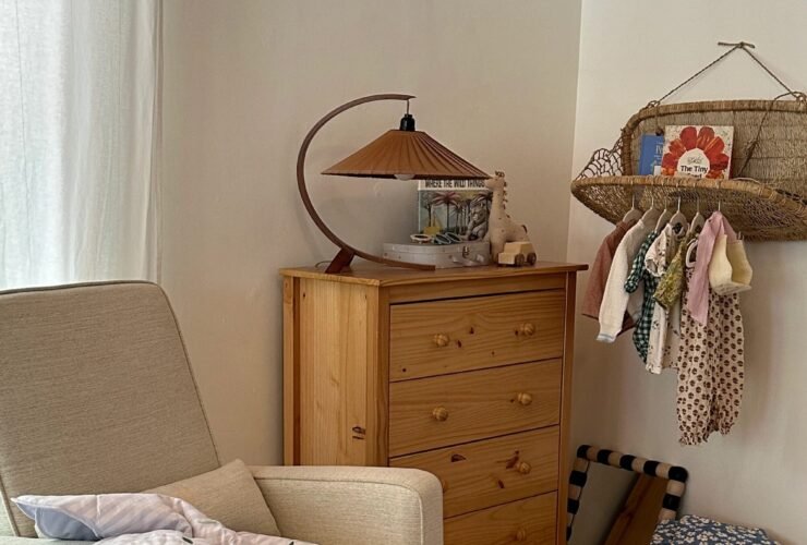 A cozy nursery with an armchair draped with a blanket, a wooden dresser with a lamp and decor, a hanging rack with baby clothes, and a patterned rug on a carpeted floor.