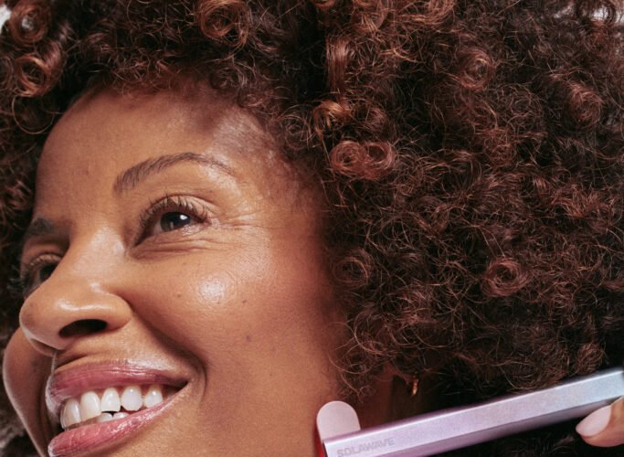 A smiling person with curly hair uses a skincare device on their chin, wearing a white top.