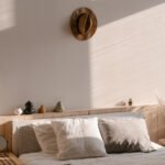 Close up shot in a bedroom of a wooden bed frame with white pillows, white sheets, and a grey comforter on top. A brown fedora hangs on the wall above the bed.
