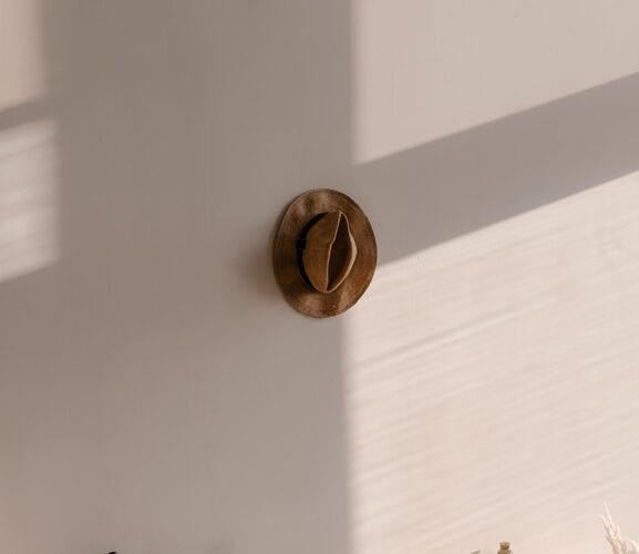 Close up shot in a bedroom of a wooden bed frame with white pillows, white sheets, and a grey comforter on top. A brown fedora hangs on the wall above the bed.