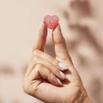 A hand with light-colored nail polish holds a small, pink heart-shaped candy with a "V" on it.