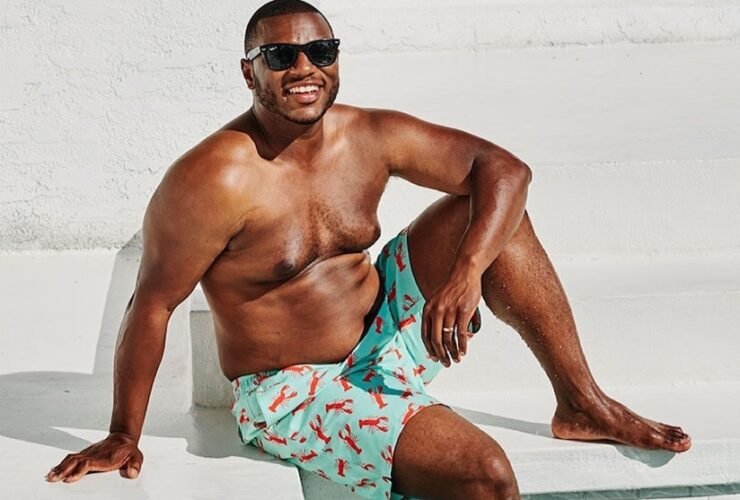A man in shorts and sunglasses sits on white steps by a swimming pool, smiling.