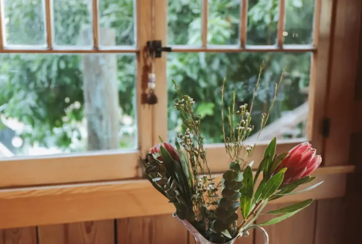 A wooden table with an open cookbook, wooden spoon, vase with flowers, a bowl of fruits, and a window with greenery outside.