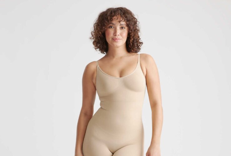 A person with curly hair is wearing a beige, form-fitting bodysuit. They are barefoot and standing against a plain white background.