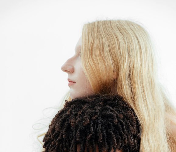A person with long blonde hair embraces another person with short, curly hair from behind, both gazing forward with neutral expressions against a plain white background.