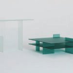 Two minimalist glass tables, one taller and clear, the other shorter and green, displayed against a plain background.