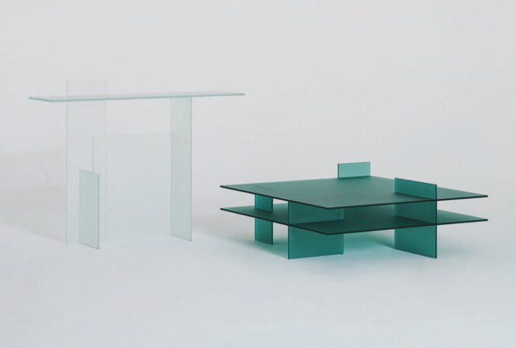 Two minimalist glass tables, one taller and clear, the other shorter and green, displayed against a plain background.