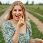 A person with long blonde hair in a green floral dress smiles and holds a strawberry to their mouth while standing in a strawberry field. They have a container of strawberries in their other hand.