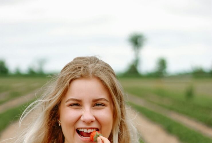 A person with long blonde hair in a green floral dress smiles and holds a strawberry to their mouth while standing in a strawberry field. They have a container of strawberries in their other hand.