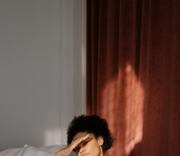 A person in a white dress sits on a couch with a laptop, shielding their eyes with one hand.