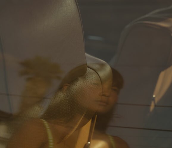 Woman in a green bikini top is seen through a car window with reflections of a palm tree and power lines.