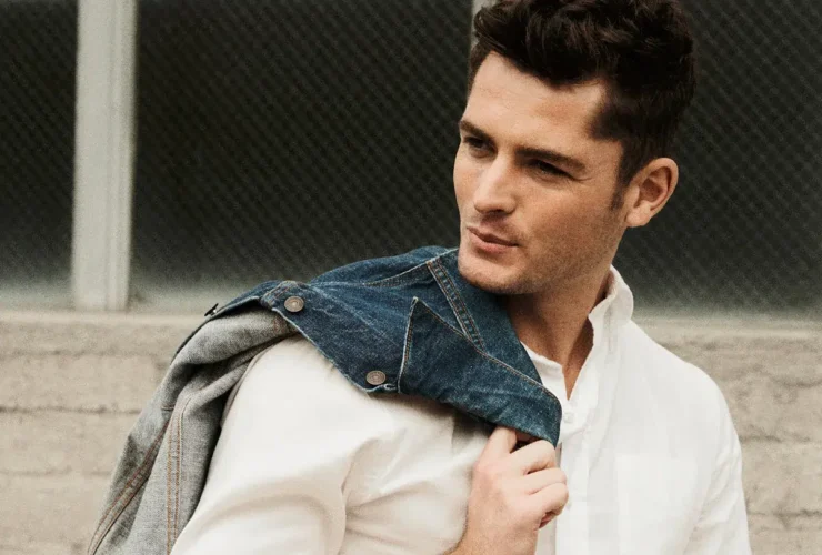 A man wearing a white shirt and grey pants holds a denim jacket over his shoulder while standing in front of a brick wall.