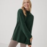 A model wearing a green Pact knit sweater and beige leggings.