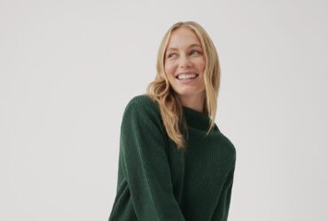 A model wearing a green Pact knit sweater and beige leggings.