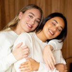 Two women embracing and smiling at the camera, one with blonde hair and the other with dark hair, both wearing white sweatshirts.