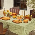 Outdoor dining table set with a green checkered tablecloth, terracotta plates, bowls, cutlery, glassware, candles, a bottle of wine, yellow flowers, and small tomatoes. Wooden chairs surround the table.