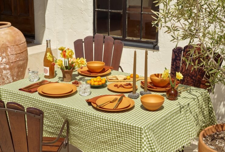 Outdoor dining table set with a green checkered tablecloth, terracotta plates, bowls, cutlery, glassware, candles, a bottle of wine, yellow flowers, and small tomatoes. Wooden chairs surround the table.