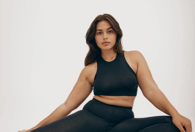 A studio shot of a model seated in pigeon pose wearing a black Organic Basics cotton leggings and high neck sports bra set.