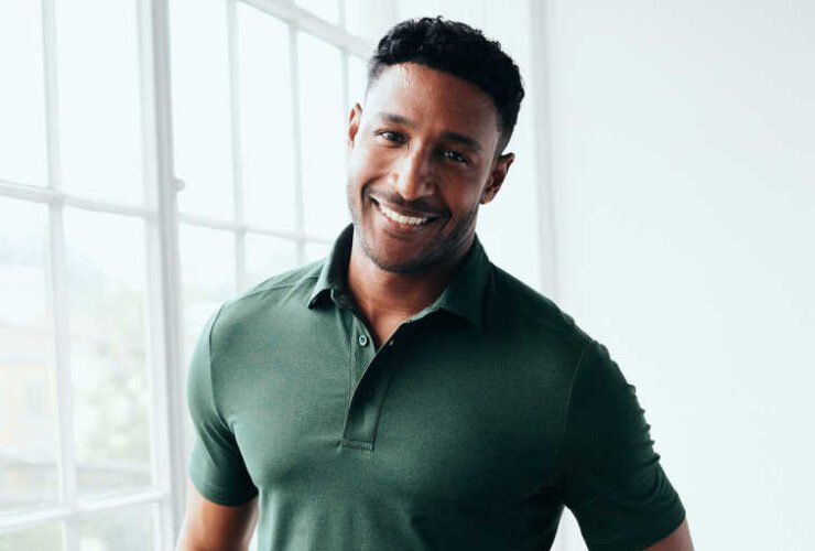 A man in a green polo shirt and black pants stands smiling by a window with natural light.