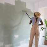 Person painting a white wall green using a roller in a brightly lit room, wearing overalls and a headscarf. Plastic sheet covers the floor below.