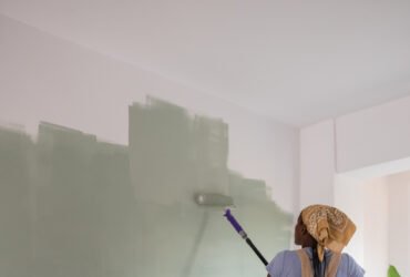 Person painting a white wall green using a roller in a brightly lit room, wearing overalls and a headscarf. Plastic sheet covers the floor below.
