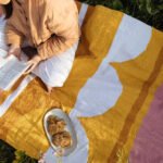 A person sits on a colorful patterned blanket outdoors, holding a book or notebook. A plate with cookies is placed on the blanket near the person. Greenery and flowers are visible around the blanket.