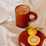 A brown mug with a frothy drink, a small orange and a lemon slice on a wooden plate with a knife, all placed on a light fabric surface.