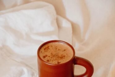 A brown mug with a frothy drink, a small orange and a lemon slice on a wooden plate with a knife, all placed on a light fabric surface.