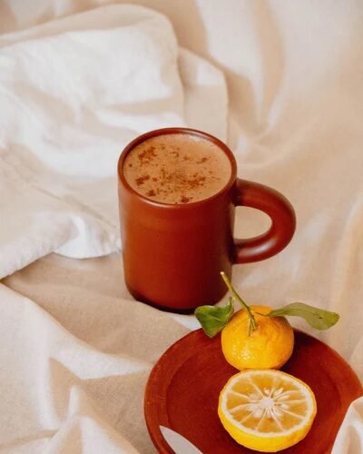 A brown mug with a frothy drink, a small orange and a lemon slice on a wooden plate with a knife, all placed on a light fabric surface.