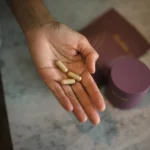 A hand holding three capsule-shaped prenatal vitamins with a blurred background showing a purple container and a similar colored pouch.