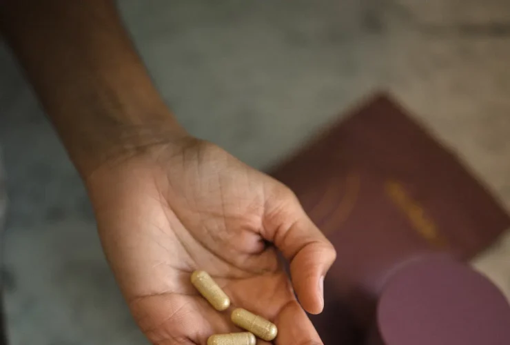 A hand holding three capsule-shaped prenatal vitamins with a blurred background showing a purple container and a similar colored pouch.