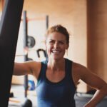 A person in a gym, wearing a blue tank top, smiles at the camera while leaning on a piece of gym equipment.