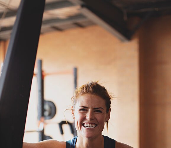 A person in a gym, wearing a blue tank top, smiles at the camera while leaning on a piece of gym equipment.