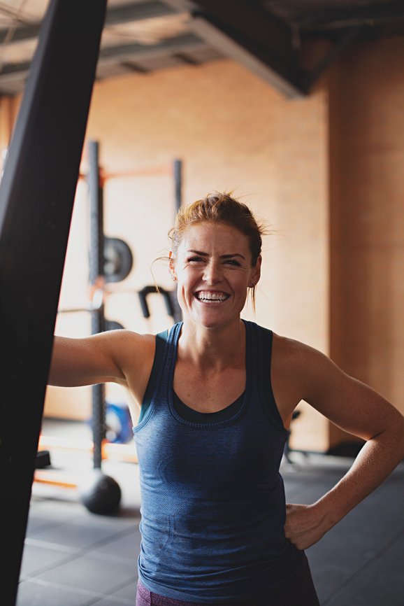 A person in a gym, wearing a blue tank top, smiles at the camera while leaning on a piece of gym equipment.