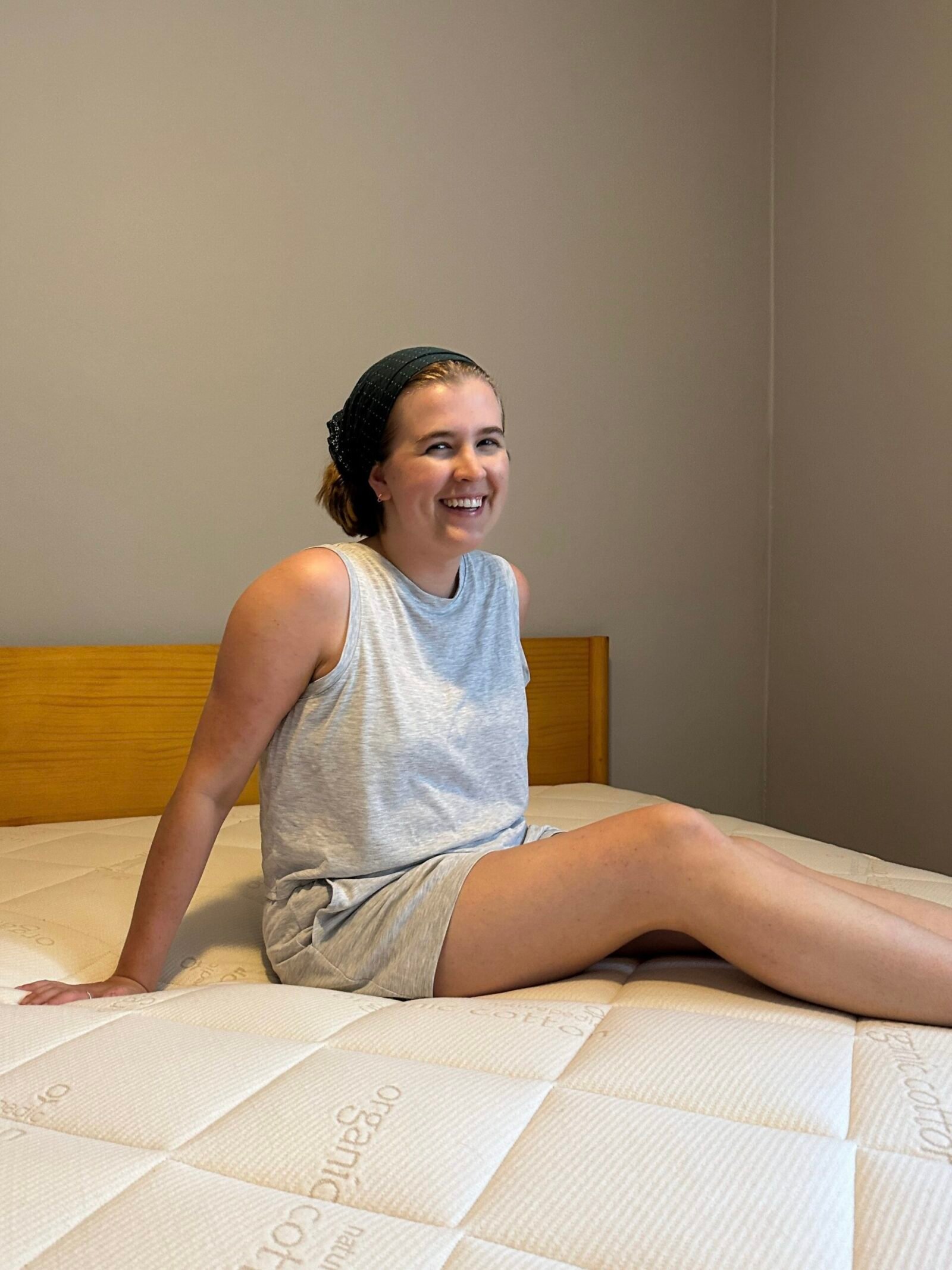 A person sits on a bed with a light grey comforter, wearing a grey tank top and shorts, and smiling. The room has plain walls and a wooden headboard.