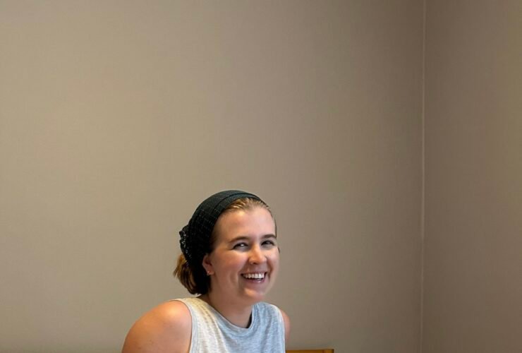 A person sits on a bed with a light grey comforter, wearing a grey tank top and shorts, and smiling. The room has plain walls and a wooden headboard.