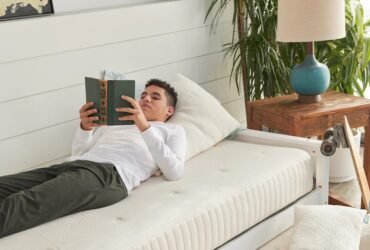 A person lies on a twin bed reading a book, with a trundle bed below, next to a wooden nightstand and a potted plant.