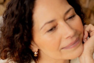 A person with curly hair wearing a beige cardigan and gold hoop earrings looks off-camera with a slight smile.