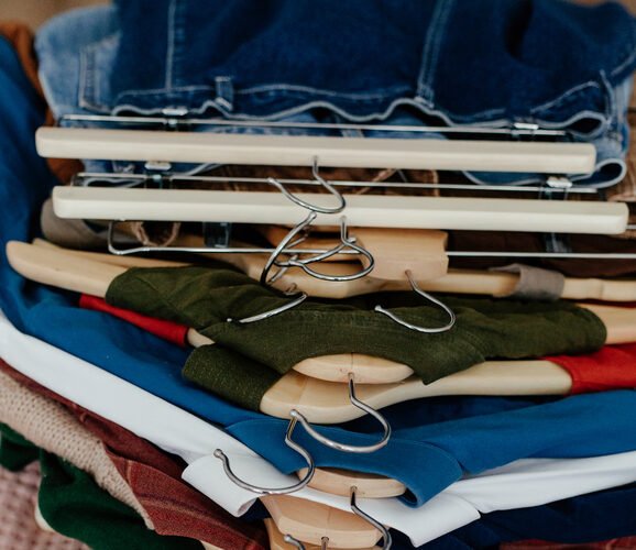 A stack of various colored clothes neatly hung on wooden and metal hangers, placed on a pink textured surface.
