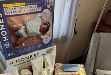 A child reaches up toward a table displaying Honest company diapers, wipes, and baby care products. An armchair is visible beside the table.